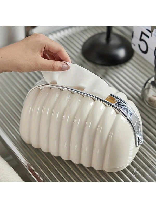 A creamy white tissue box holder with a unique design, resembling a bag, placed on a metallic surface with a person holding a tissue.