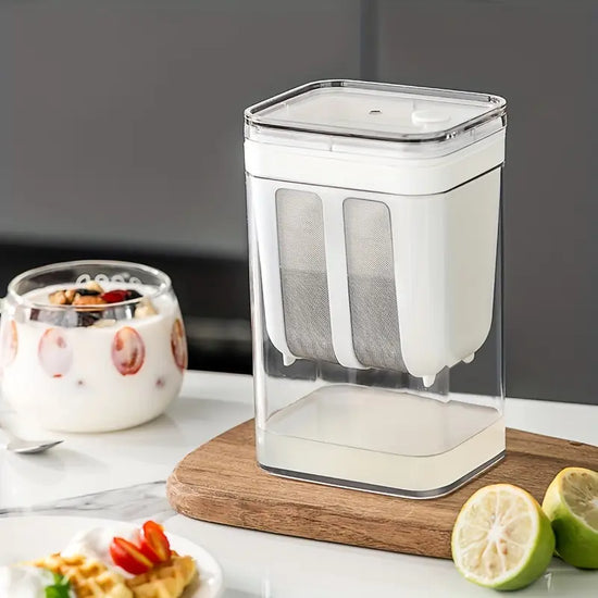 Clear kitchen storage container with white base on a wooden cutting board, with food items in the background.