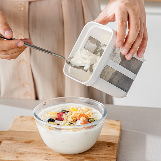 Person using a yogurt maker with a bowl of yogurt and fruit on a wooden board.